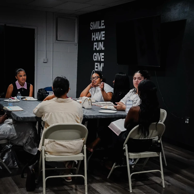 A group of women sit around a long table in a dimly lit room, engaged in a discussion. One woman gestures as she speaks. A dark wall has motivational text.