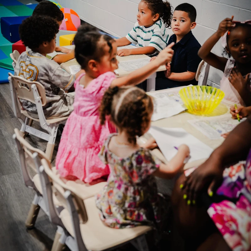 A group of diverse young children sitting at a table with paper and crayons, looking in different directions during a classroom activity.