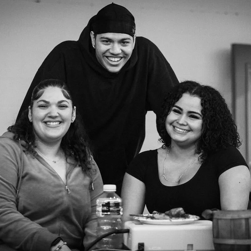 A black and white photo of three young people. Two women are seated and smiling, while a man stands smiling behind them, creating a happy group portrait.