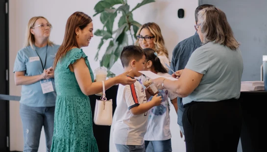 A smiling woman in a teal dress with her two young children interacts with a friendly greeter at an indoor event's check-in table.