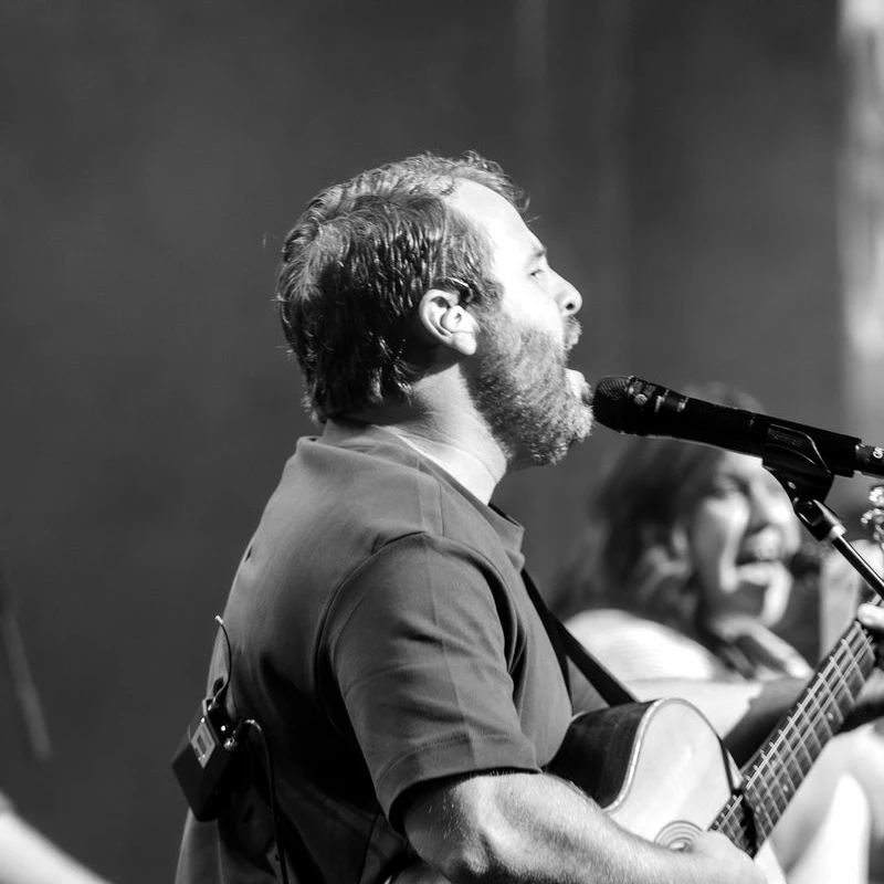A black and white side-profile photo of a bearded man singing into a microphone while playing an acoustic guitar on stage during a performance.