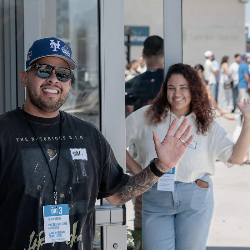 A smiling man in a baseball cap and a woman waving stand at an entrance, welcoming people to an outdoor event. They are both wearing lanyards.