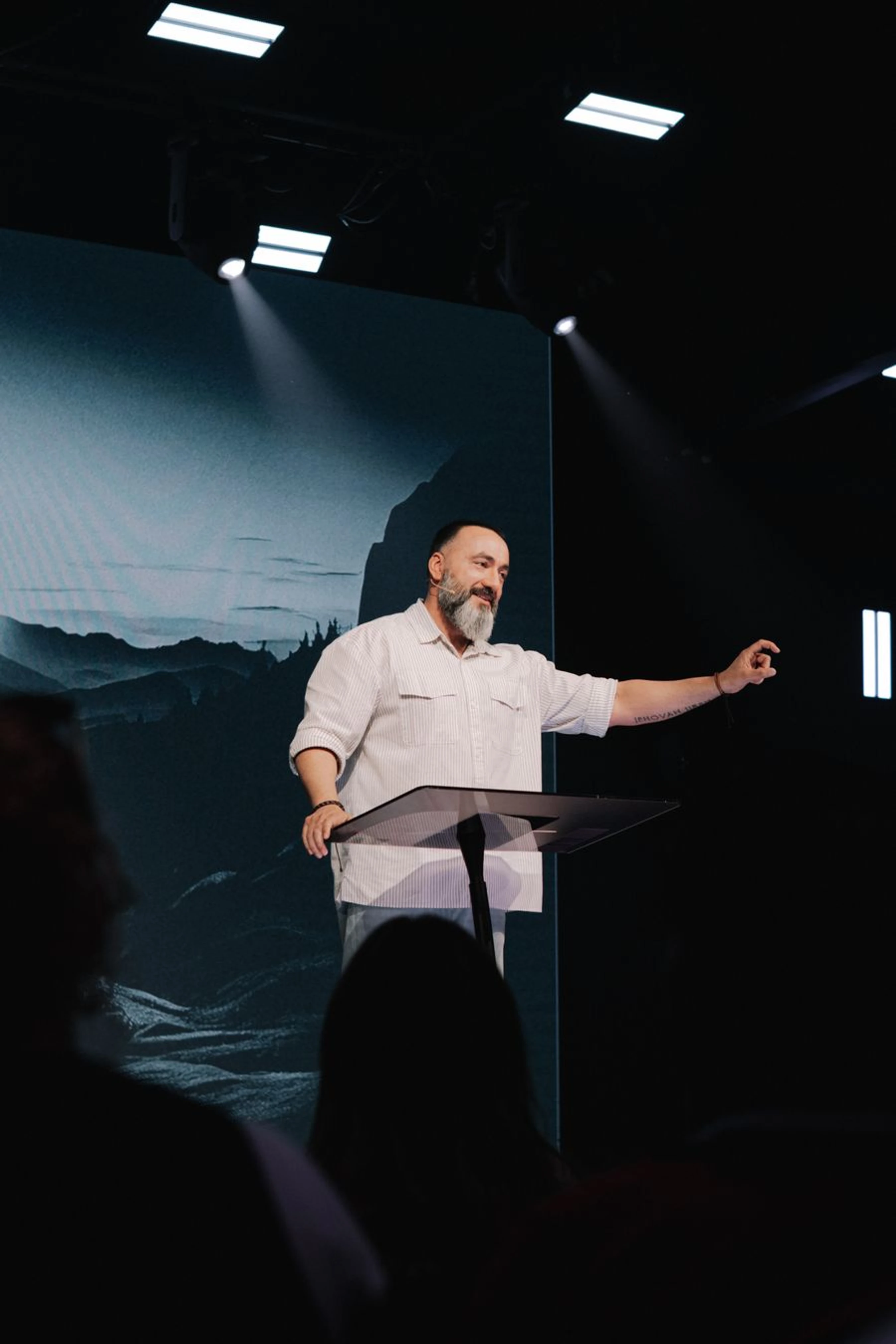 A man with a gray beard in a white shirt stands at a lectern on a dark stage, gesturing as he speaks. A large screen behind him shows a mountain landscape.
