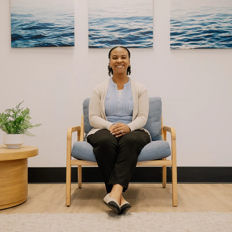 A smiling Black woman sits in a light wood armchair with blue cushions in a modern office, with a triptych of ocean photos on the wall behind her.
