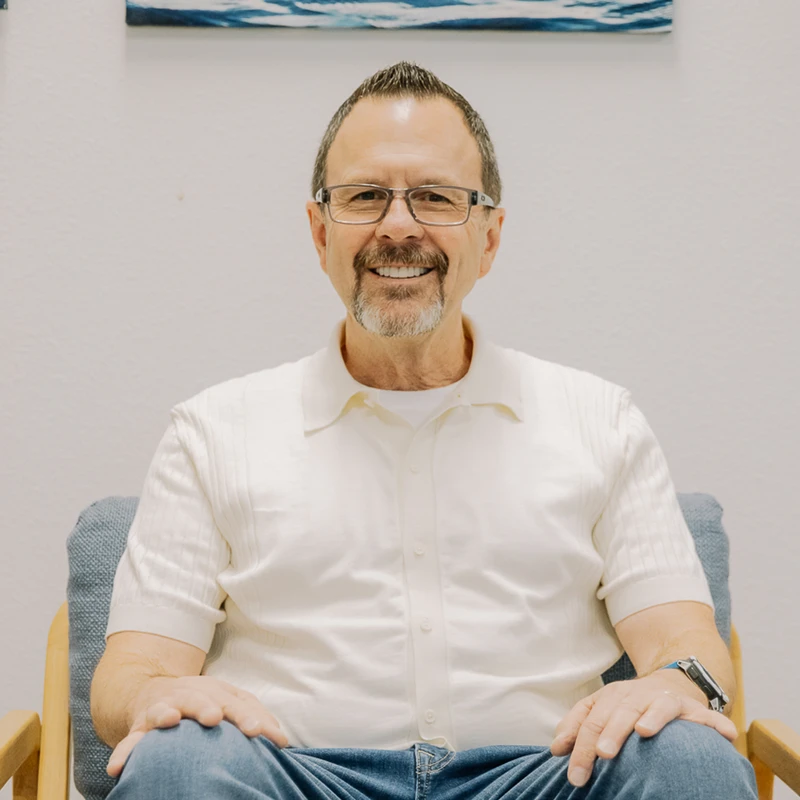 A middle-aged man with glasses and a goatee sits in a blue chair, smiling at the camera. He is wearing a white collared shirt and blue jeans.