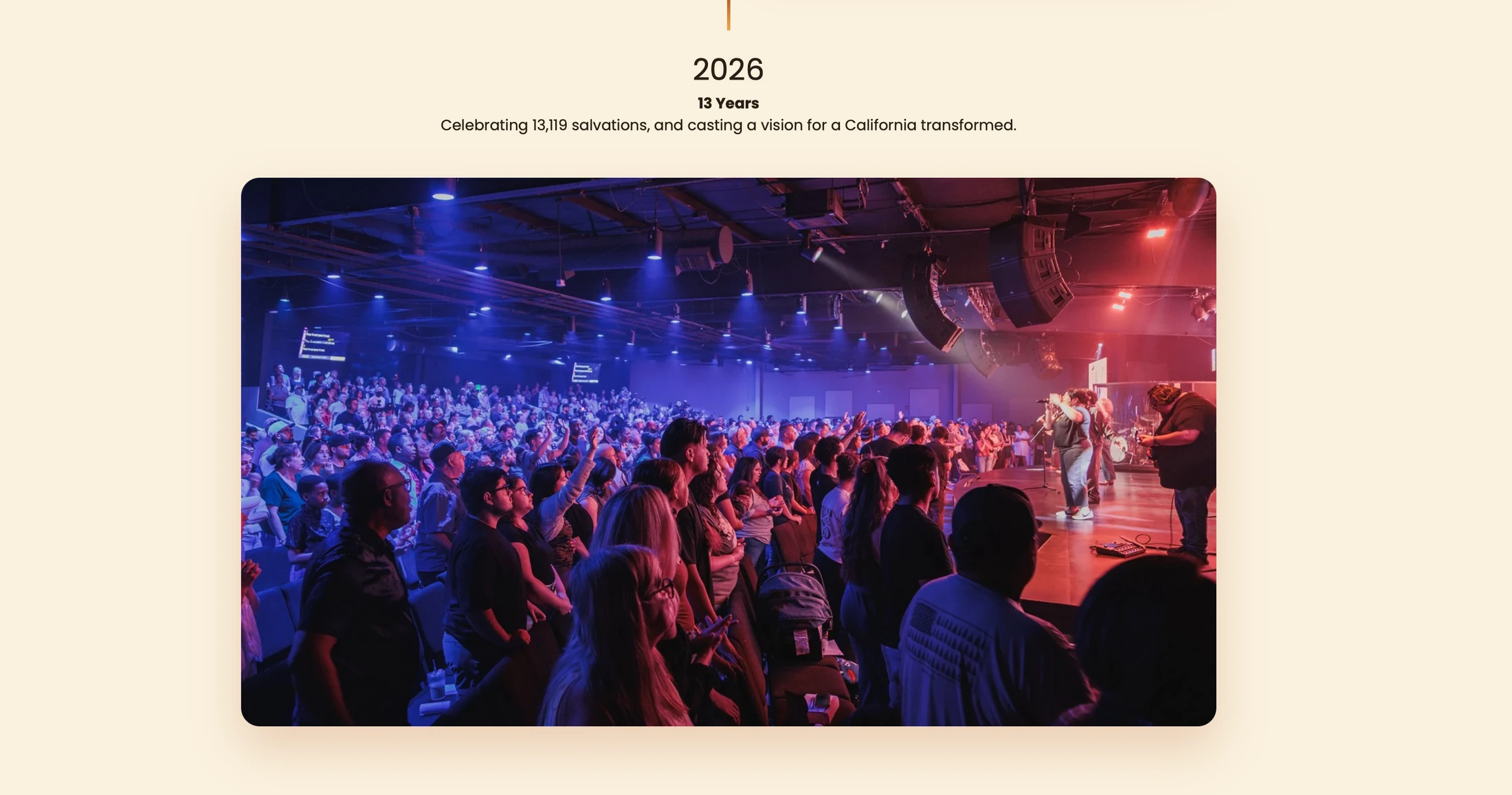 A large, diverse congregation worships during a live service. They face a band on stage, illuminated by vibrant blue and red stage lighting.