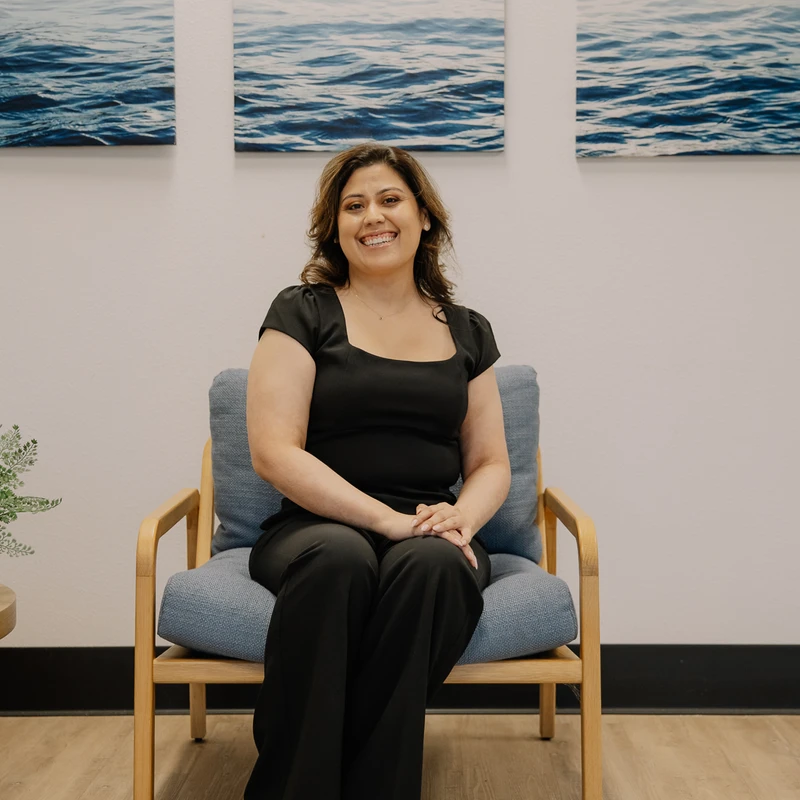 A woman in a black outfit sits smiling in a blue cushioned chair. Behind her are three pictures of ocean waves, and to her side is a table with a plant.