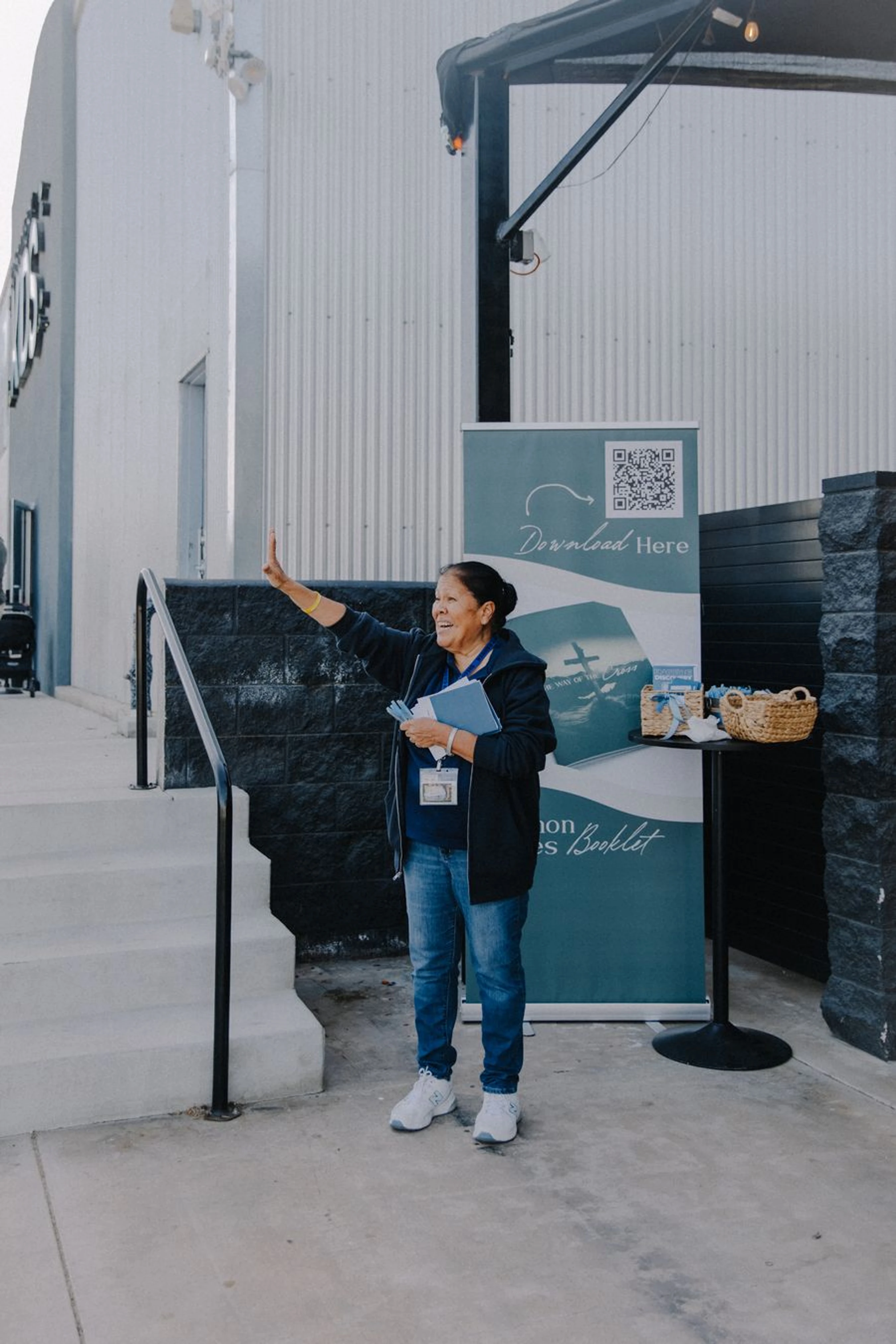 A smiling woman stands outside, waving a friendly hello. She holds blue pamphlets and wears a lanyard while standing next to an event banner with a QR code.