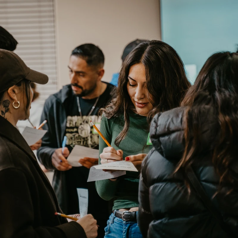 A diverse group of people stand together indoors, focused on writing on small papers with pencils during a workshop or event.
