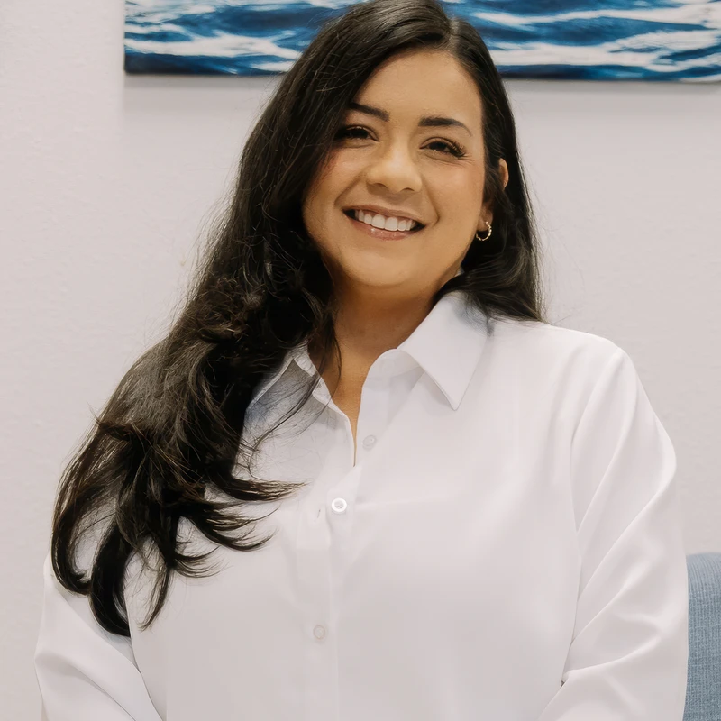 A professional headshot of a woman with long dark hair smiling at the camera. She wears a white button-up shirt and sits in front of a blue painting.