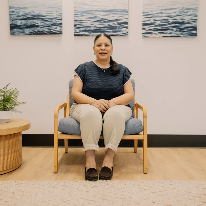 A woman with dark hair in a ponytail sits in a wooden armchair in a waiting room. She wears a navy top and looks at the camera. Ocean art hangs on the wall.