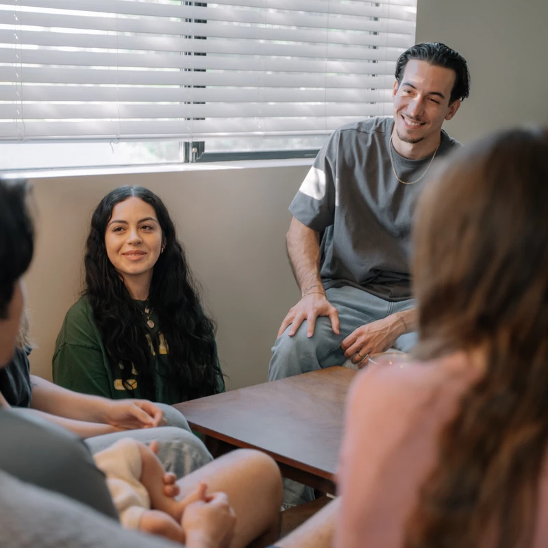 A group of four young adults sits in a circle, engaged in a conversation. A man and a woman in the background are smiling, lit by a window behind them.
