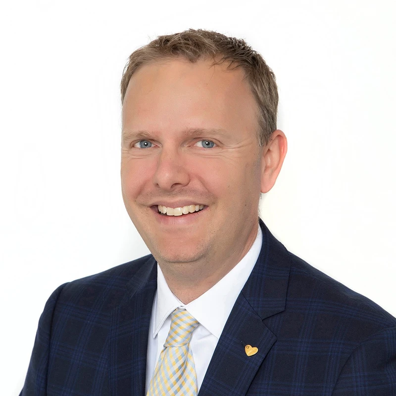 A head and shoulders portrait of a smiling white man with blue eyes and light brown hair, wearing a blue plaid suit, white shirt, and a yellow patterned tie.
