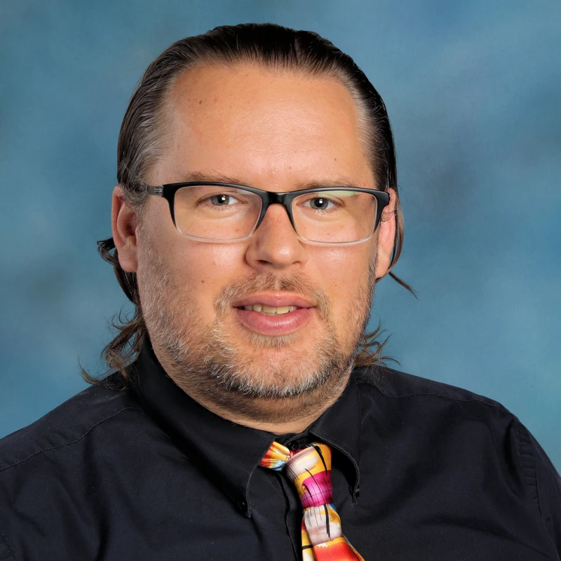 A studio headshot of a man with medium-length brown hair and glasses. He wears a black shirt and a vibrant, multicolored tie, smiling at the camera.