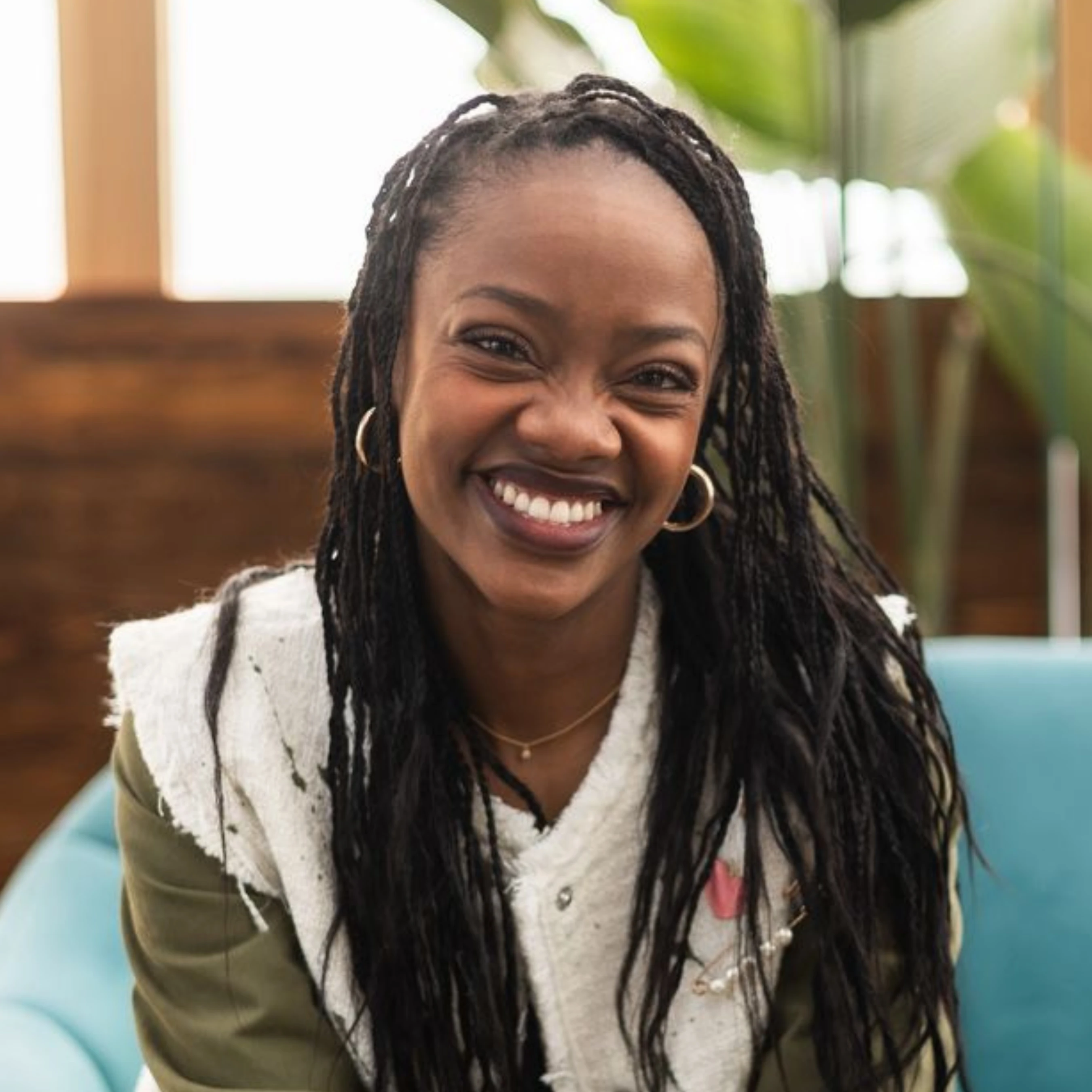 A close-up headshot of a young Black woman with long braids. She is smiling broadly at the camera while sitting on a light blue couch indoors.