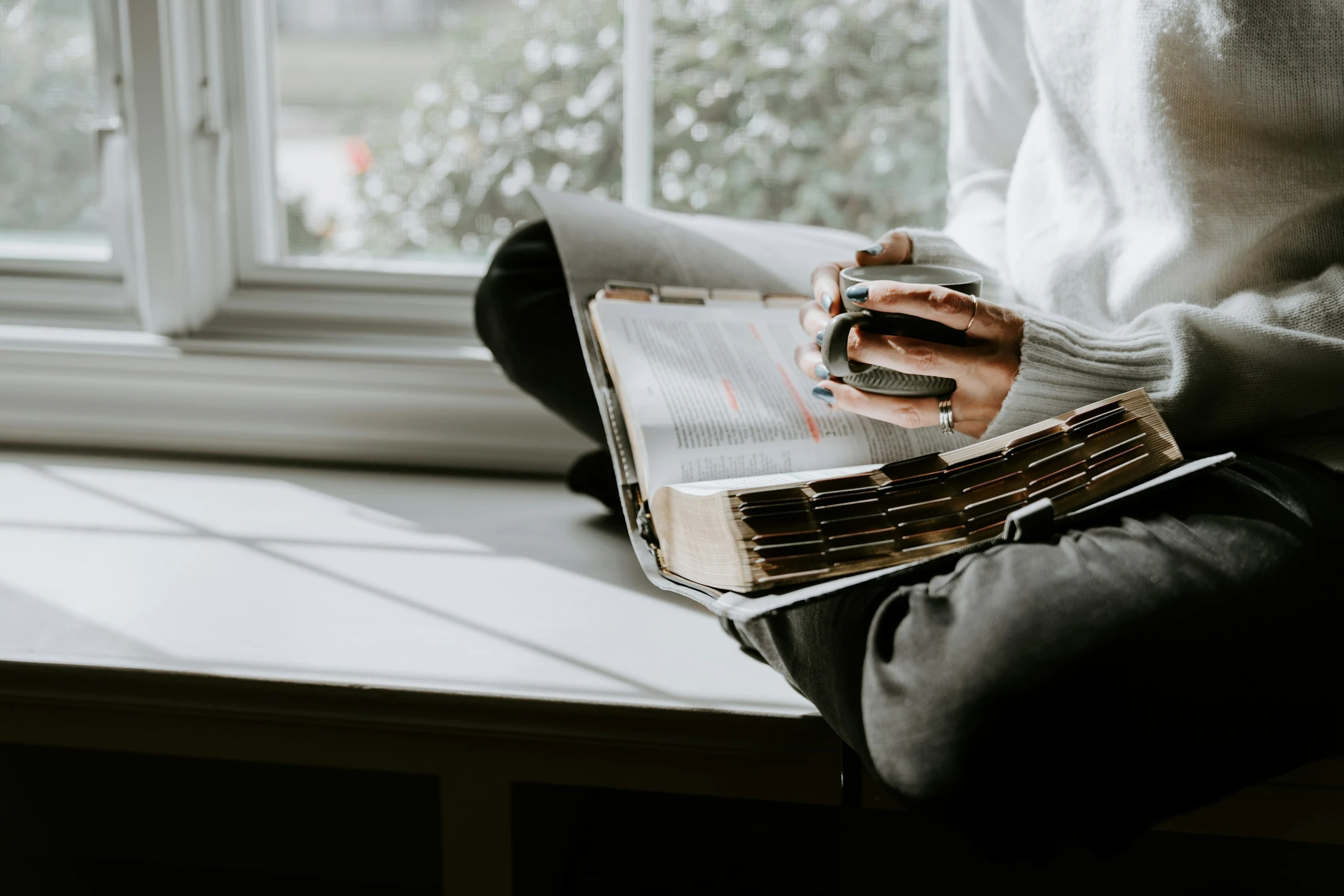 Person reading an open Bible while holding a cup of coffee by a window.