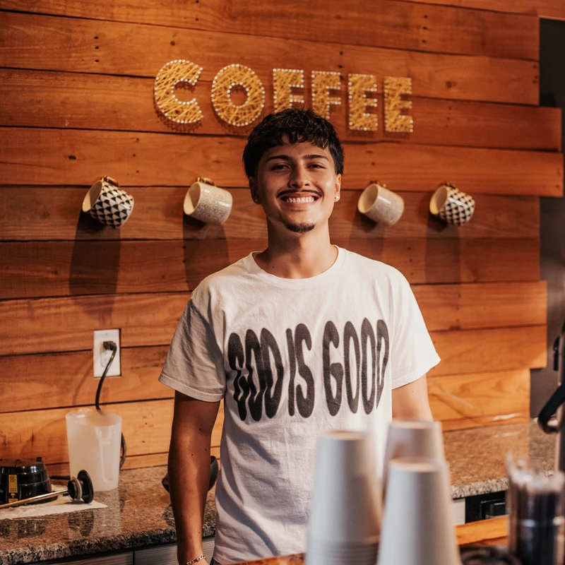 A young male barista with a mustache smiles warmly behind a counter. He wears a 'God is Good' t-shirt in front of a wooden wall with a 'COFFEE' sign.