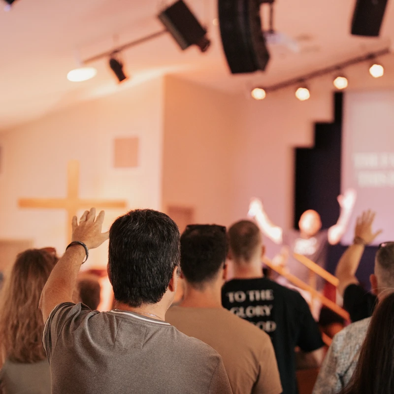 From behind, a congregation in a warmly lit church stands with their hands raised in worship, facing a stage with a screen and a wooden cross on the wall.