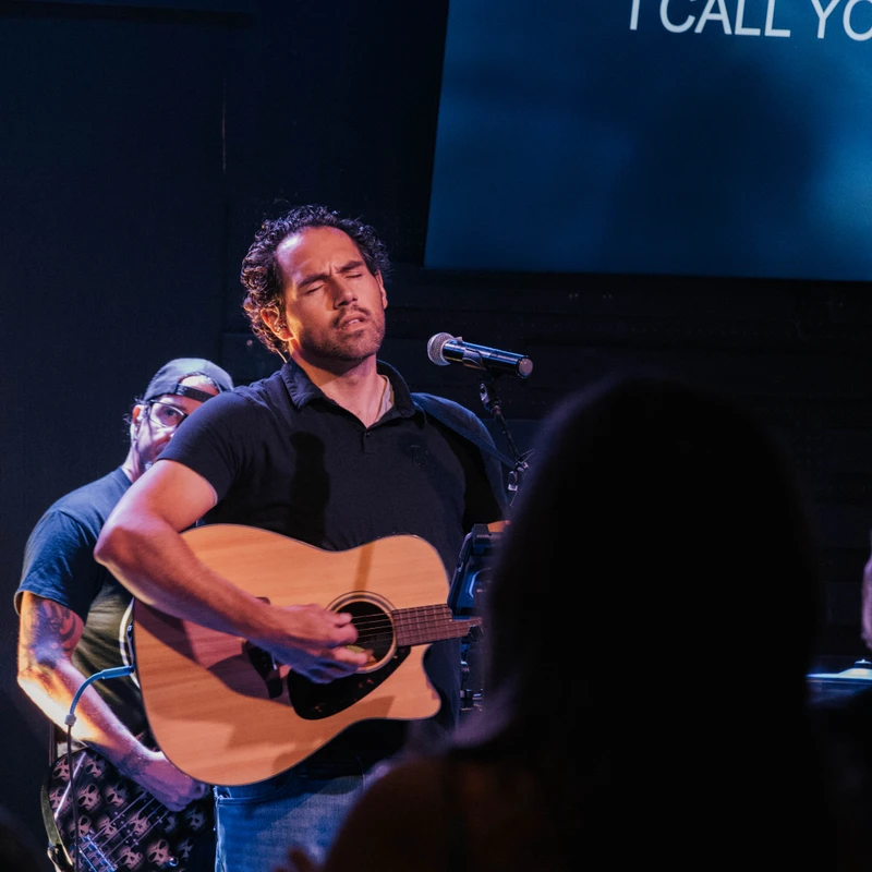 A man with dark curly hair sings with his eyes closed, playing an acoustic guitar on a dimly lit stage. Behind him, lyrics are displayed on a screen.