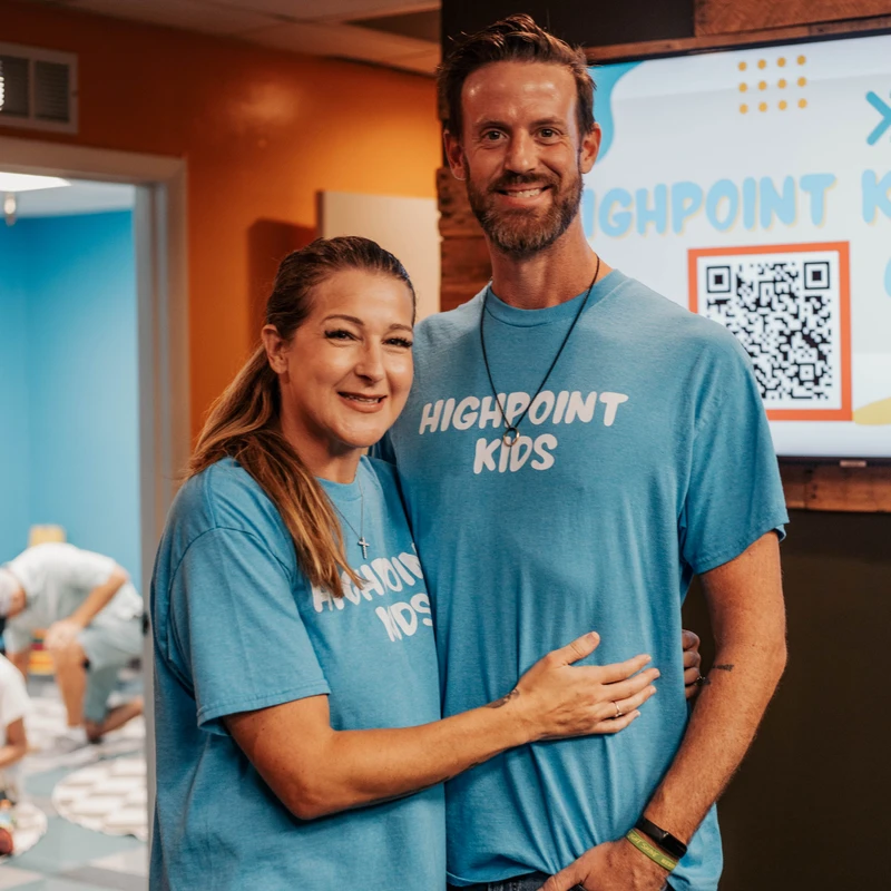 A smiling man and woman wearing matching light blue 'Highpoint Kids' t-shirts pose for a photo in a colorful room. The woman has her arm around the man.