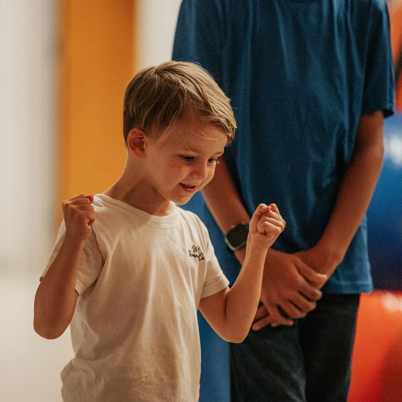 A young boy with blond hair in a white t-shirt smiles, clenching his fists in a happy, celebratory gesture while standing indoors.