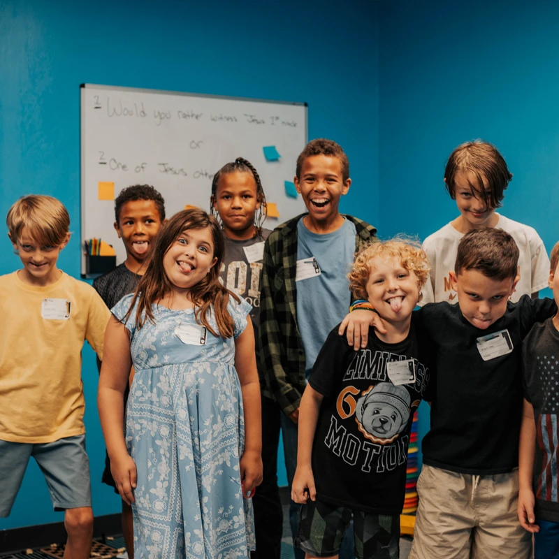 A diverse group of children stand together in a classroom with a blue wall. They are smiling and making silly faces, sticking their tongues out at the camera.