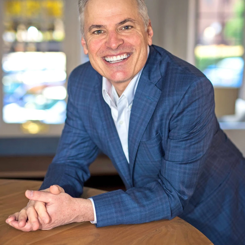 A smiling, middle-aged man with short gray hair in a blue plaid blazer and white shirt leans on a wooden table with his hands clasped, looking at the camera.