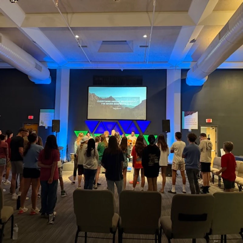 A large group of young people stand facing a stage with a projector screen during a service or meeting in a modern auditorium with blue accent lighting.