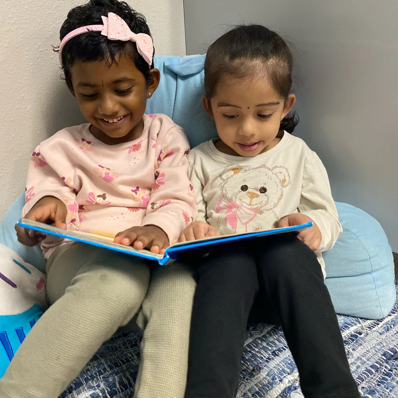 Two young girls sit close together on a blue rug, sharing a book. The girl on the left smiles happily, while the girl on the right looks down at the pages.