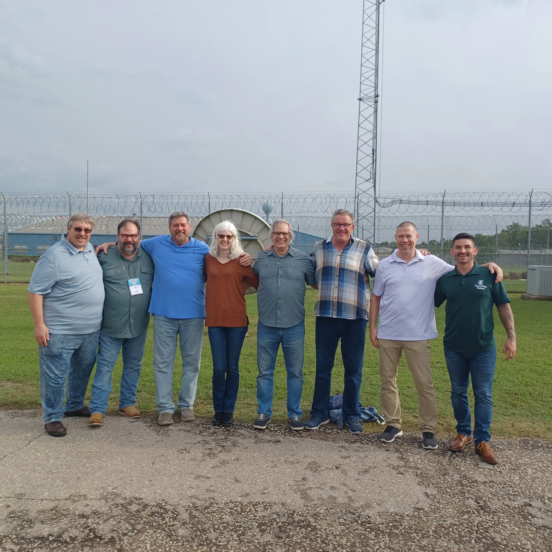 A group of eight people, seven men and one woman, stand arm in arm smiling. They are outside in front of a tall fence topped with razor wire.