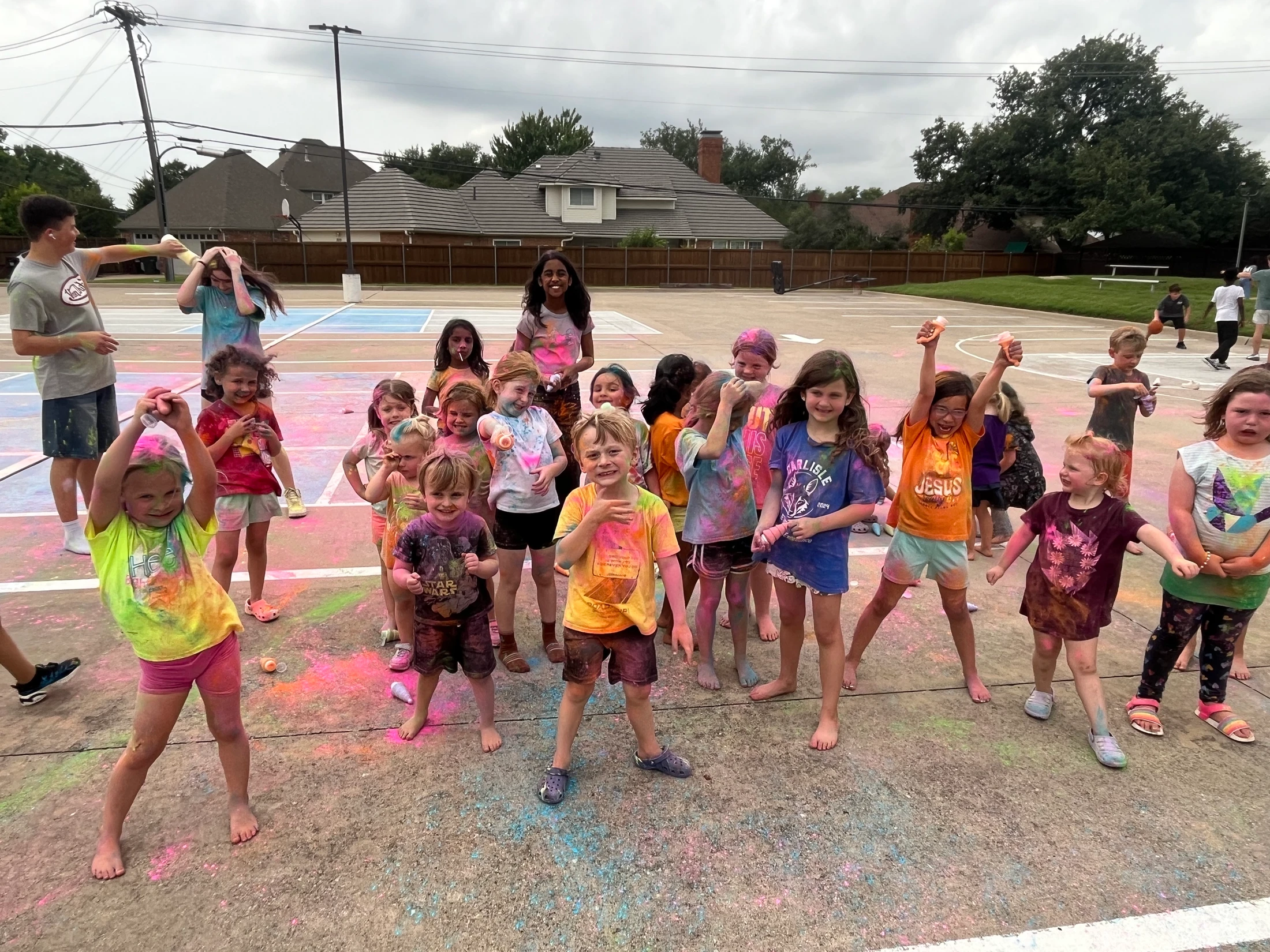 A large group of happy children and a few adults pose for a picture outdoors, all covered in multi-colored powder from a fun color run event.