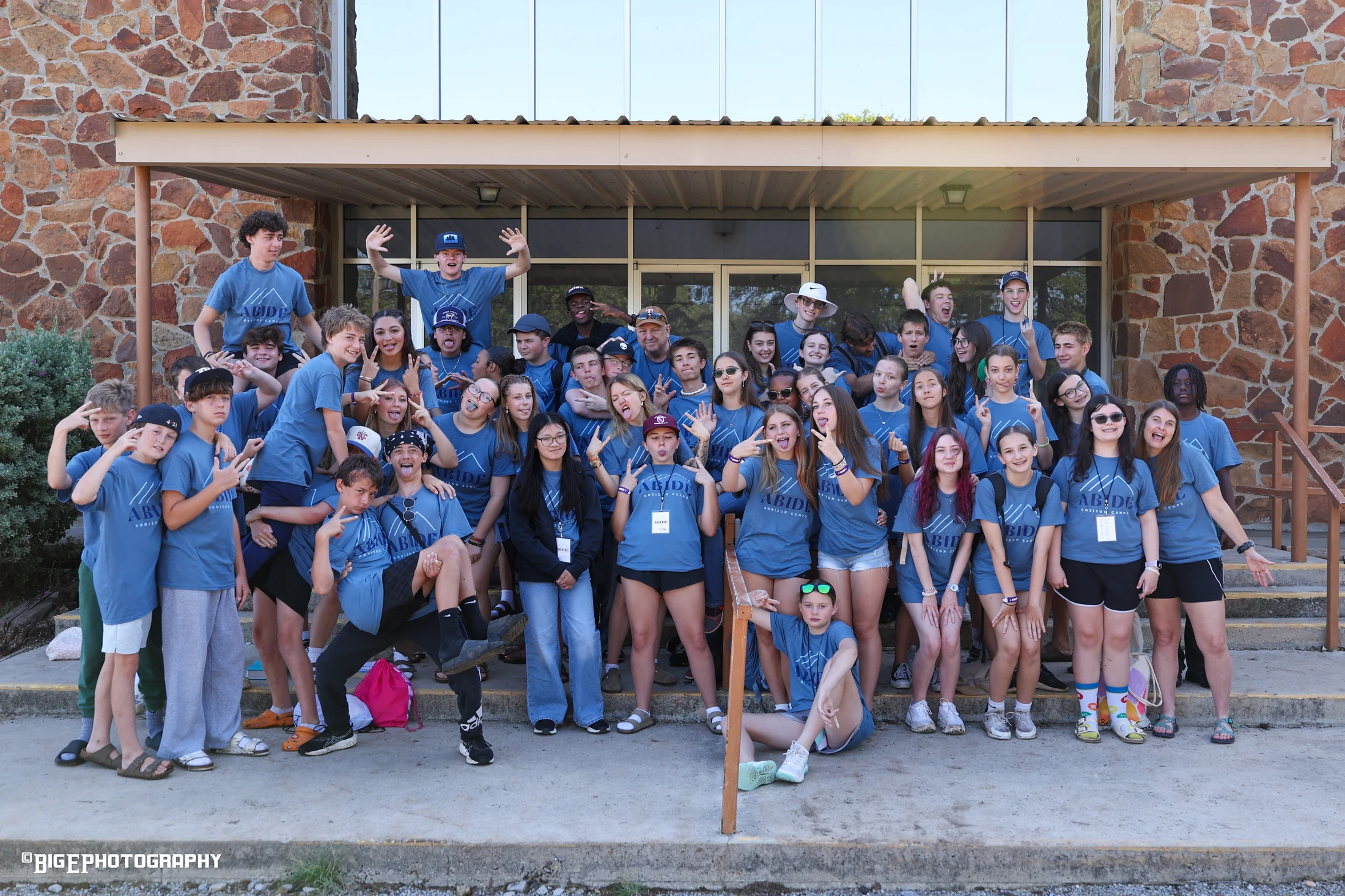 A large group of teenagers in matching blue camp t-shirts posing playfully for a group photo on the steps of a stone building.