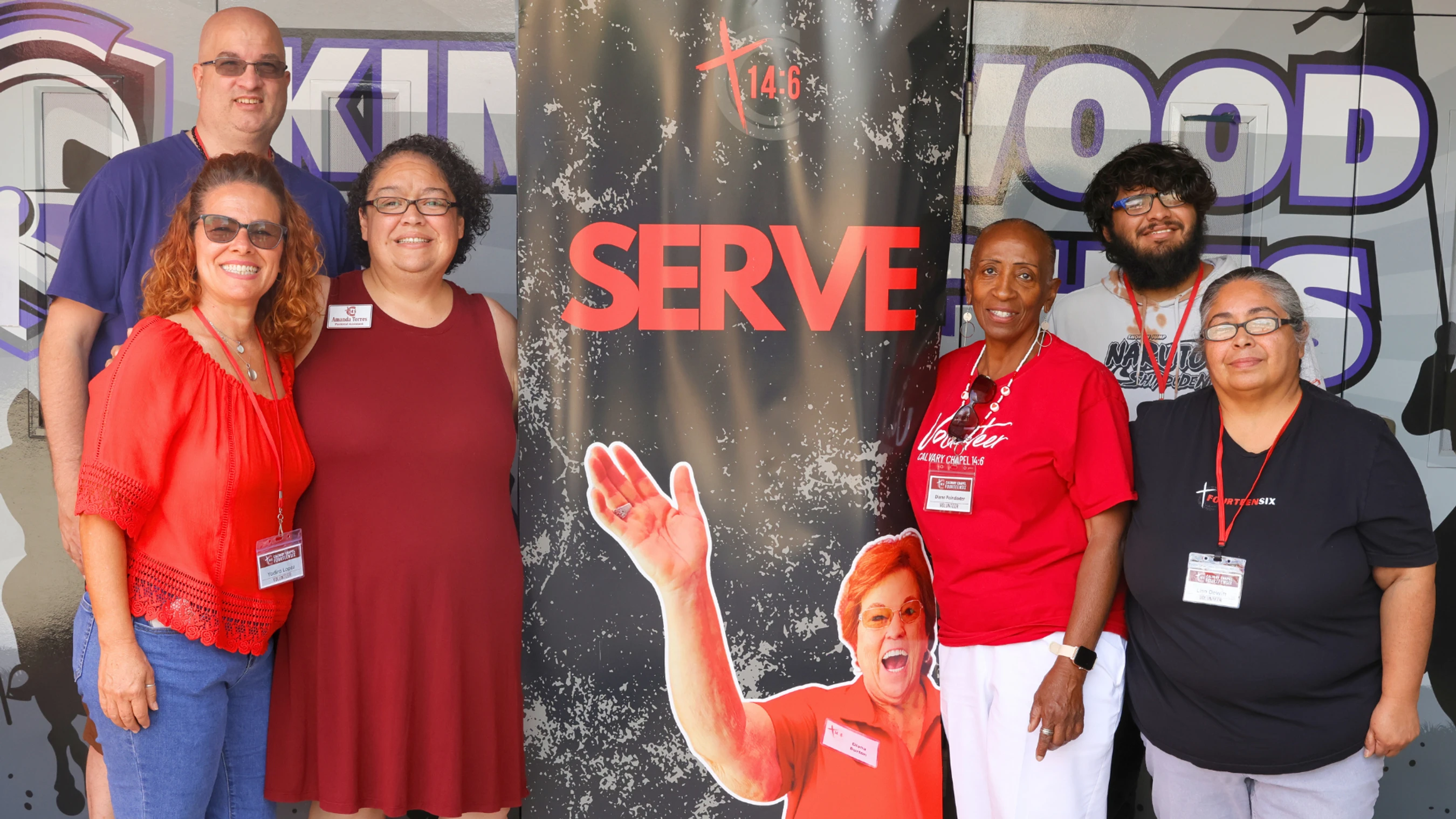 A diverse group of six adult volunteers smile together for a photo, standing in front of a black banner with the word 'SERVE' in large red letters.
