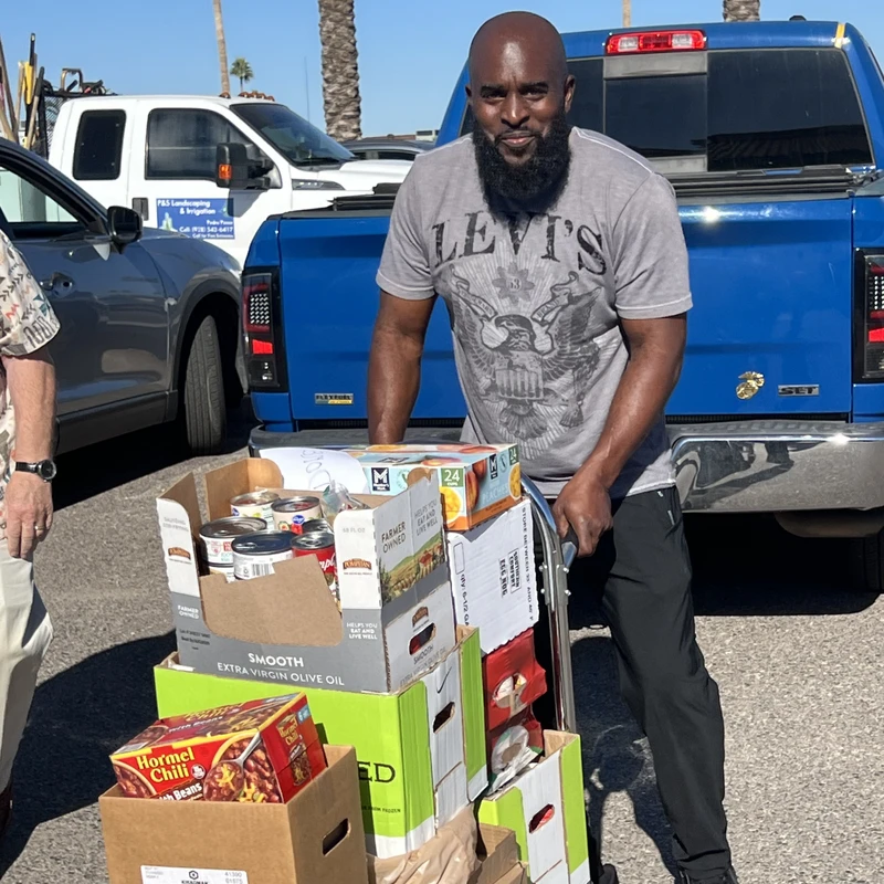 A man with a beard smiles as he pushes a hand truck stacked with boxes of donated canned goods and food items in a sunny parking lot.