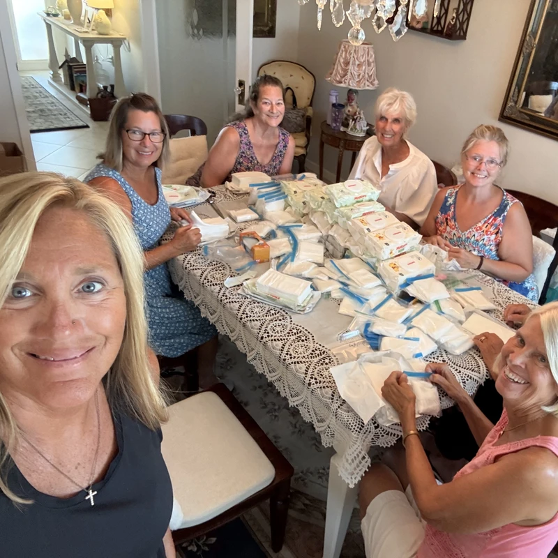 A selfie of six smiling women gathered around a dining table covered with baby wipes and other supplies, assembling them into small packages for charity.