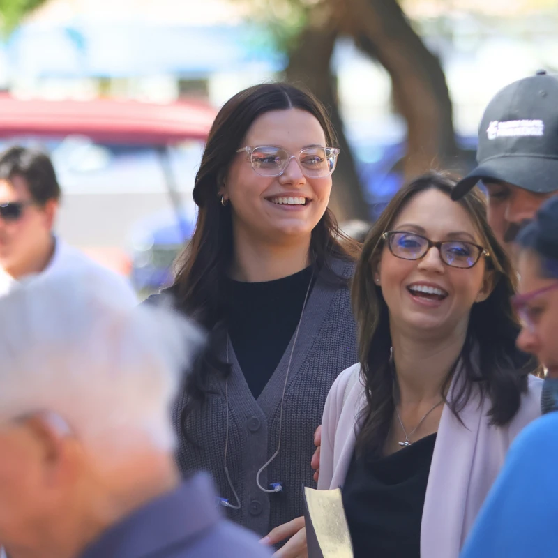 A group of people are gathered outside on a sunny day. Two women in the center are the focus, both smiling and wearing glasses in a candid moment.