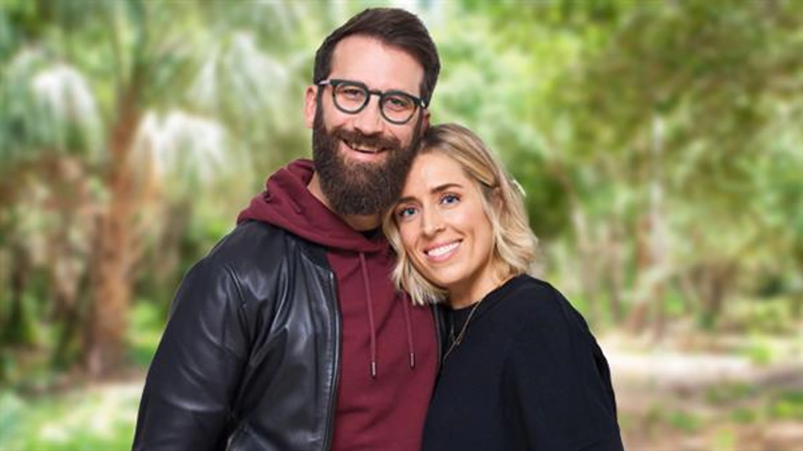 A man with a beard and glasses hugs a woman with blonde hair. They are both smiling at the camera with a blurry green, outdoor background.