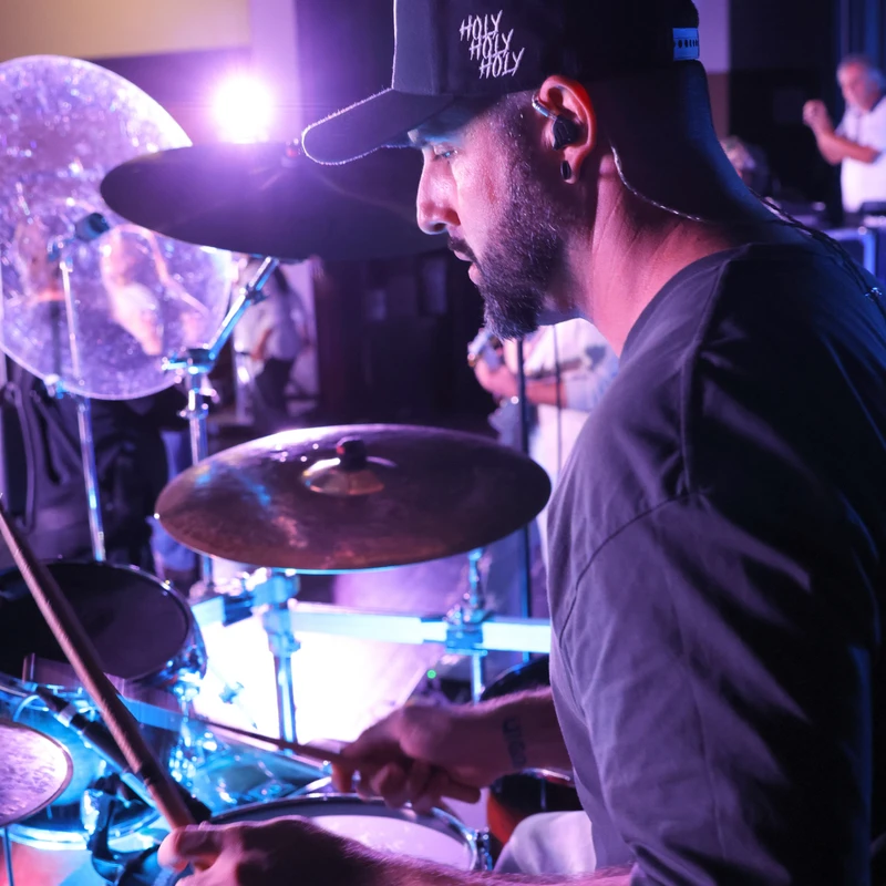 Profile view of a focused drummer wearing a cap and in-ear monitors, playing his drum kit during a performance with purple stage lighting.