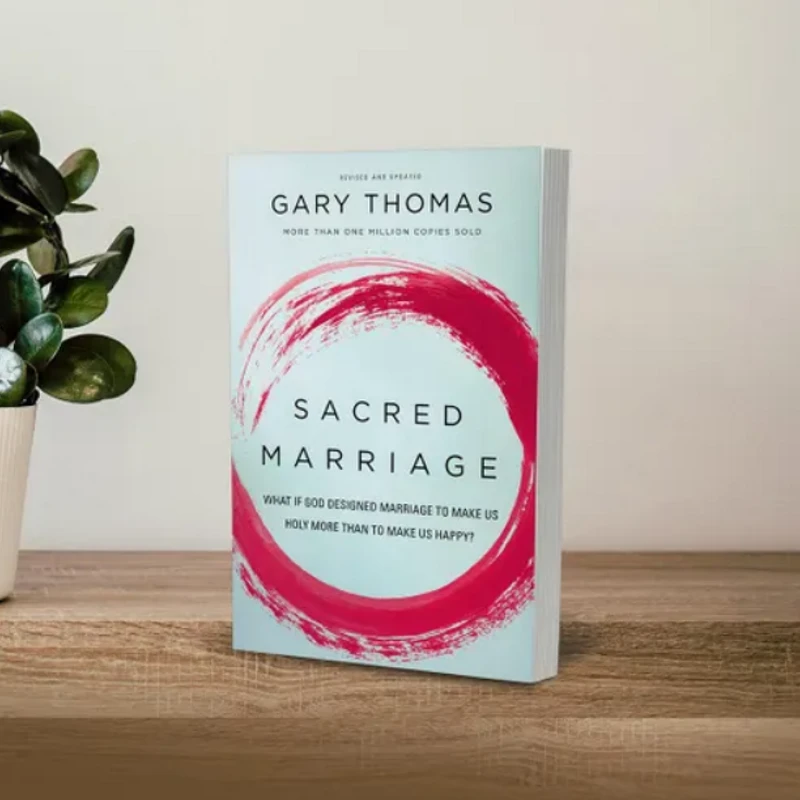A copy of the book 'Sacred Marriage' stands on a wooden shelf next to a small potted plant with green leaves against a plain wall.