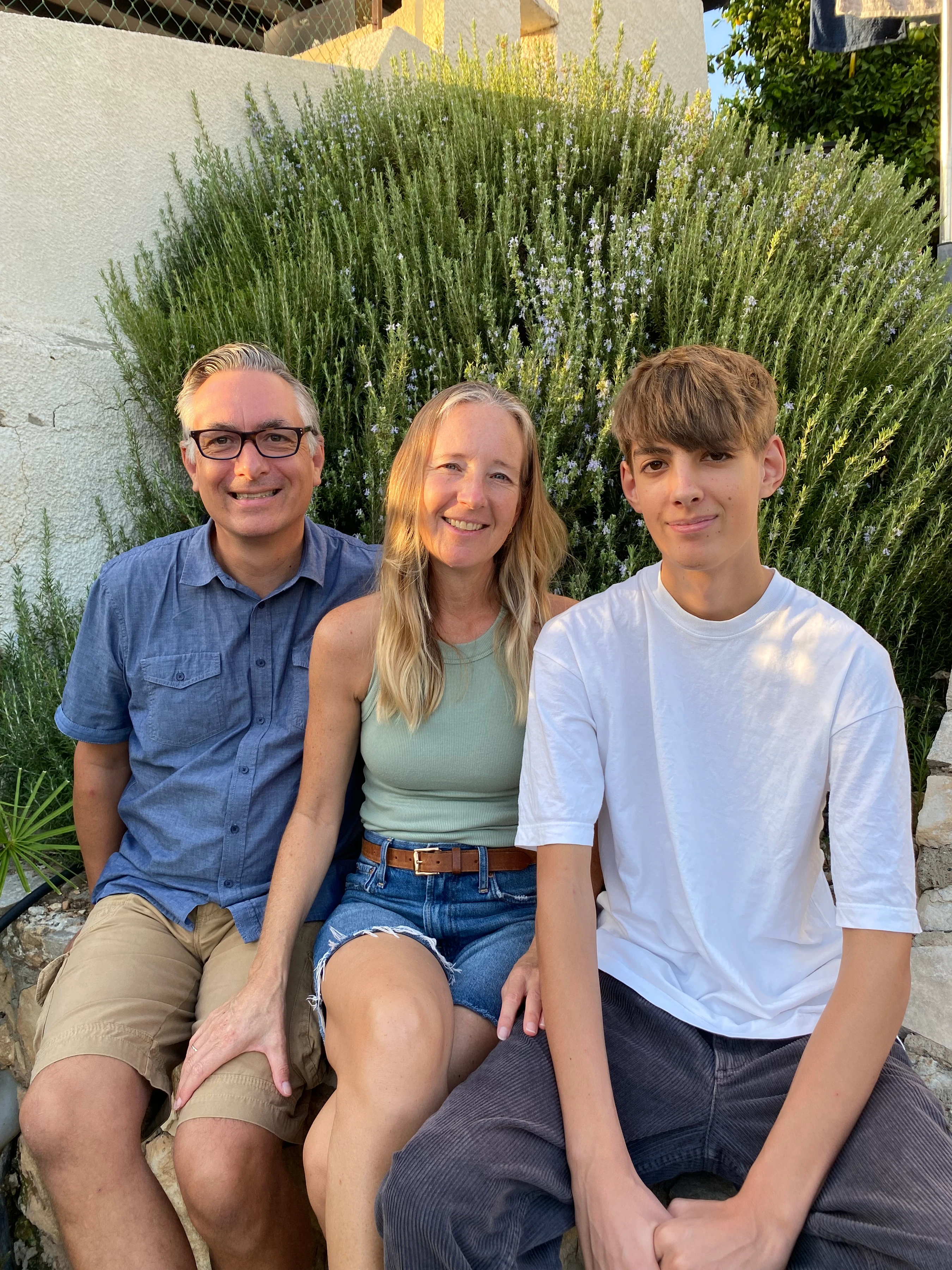 A smiling family of three, a mother, father, and teenage son, sit close together on a stone bench outdoors in front of a large rosemary bush.