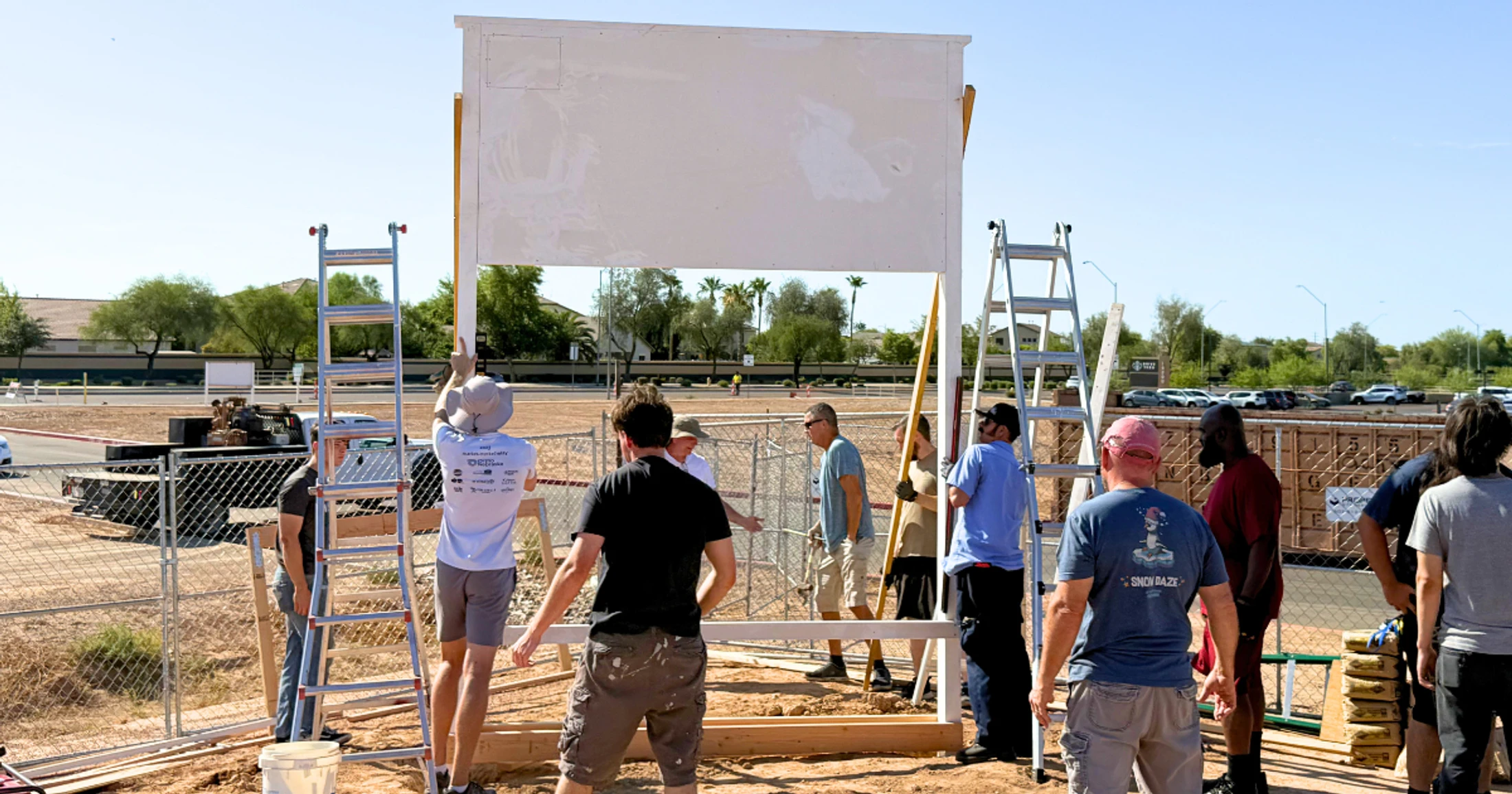 A group of people work together on a sunny day, lifting a large white panel onto a wooden frame at a construction site, with ladders and tools nearby.