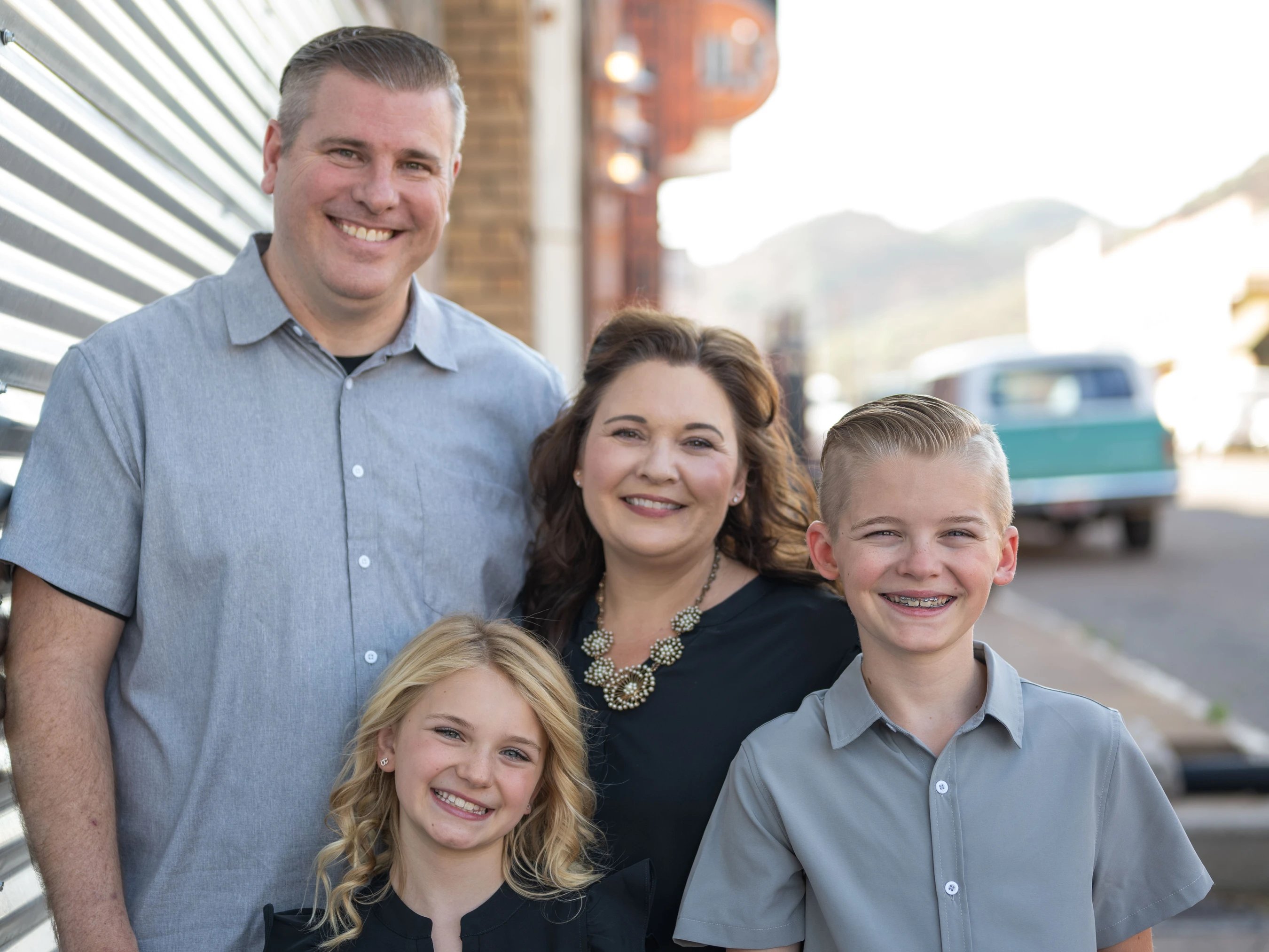 A smiling family of four, with parents standing behind their son and daughter, pose for a portrait on an urban street.