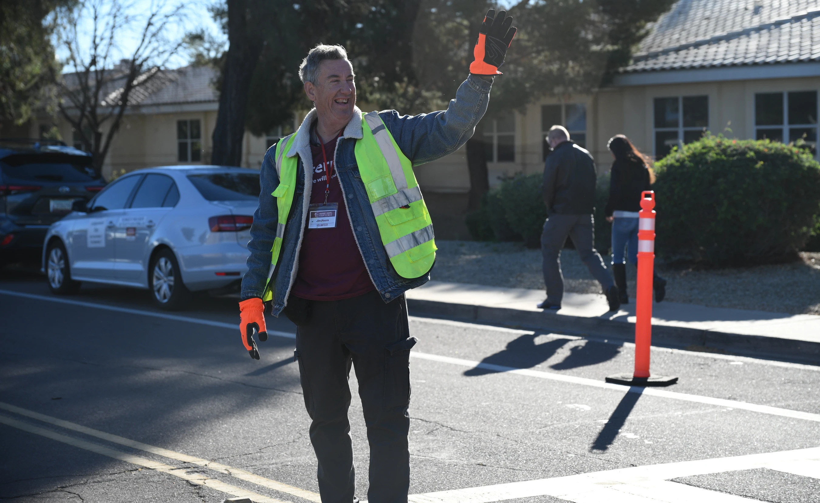 A smiling man in a yellow safety vest and orange gloves stands in a street, waving his hand to direct traffic or greet people during an outdoor event.