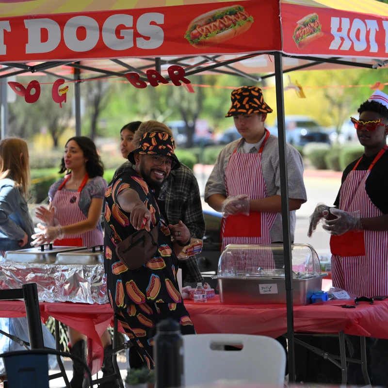 A man in a hot dog print shirt smiles and points at an outdoor hot dog stand, where several people in aprons are serving food under a red tent.