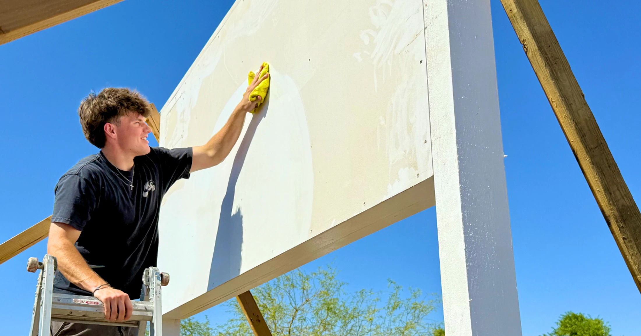 A young man on a ladder smiles as he wipes down a large white wall with a yellow cloth under a clear blue sky, preparing it for painting.