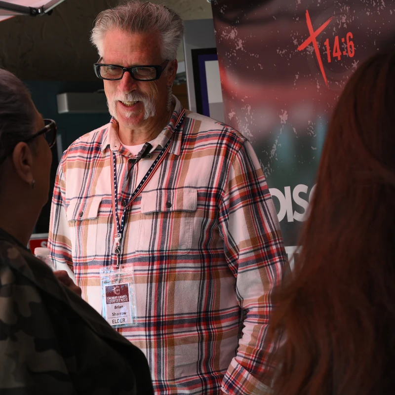 A man with gray hair, glasses, and a mustache wears a plaid shirt and a lanyard while talking with two women, seen from behind, under a canopy.