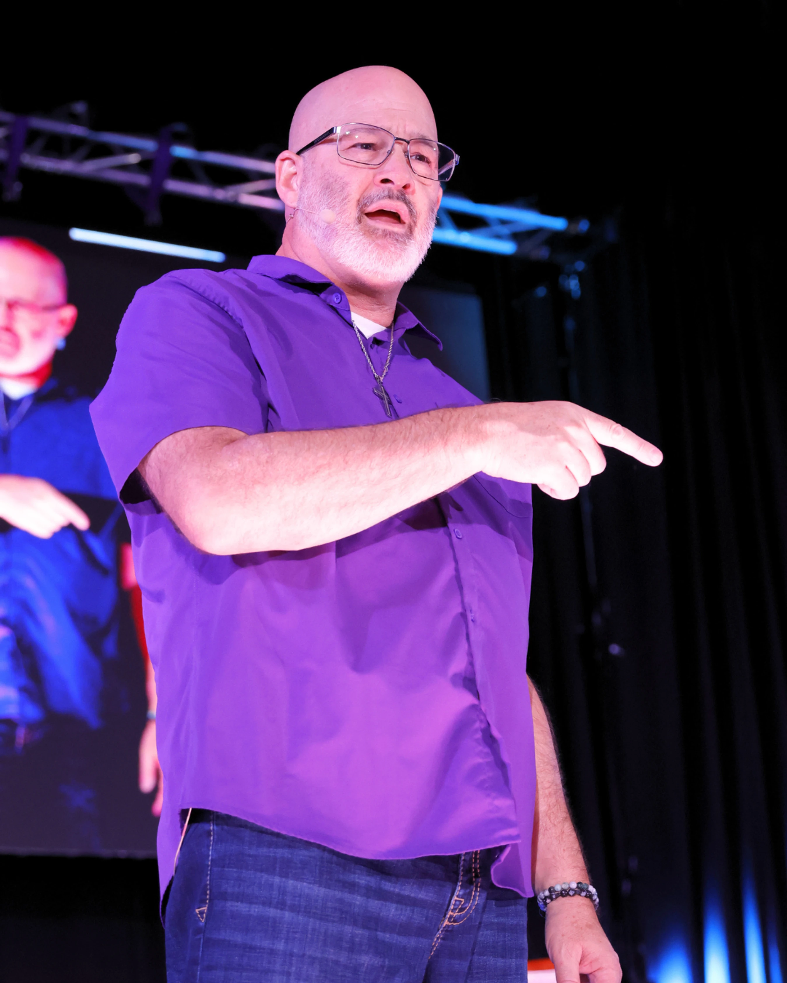 A bald man with glasses and a goatee, wearing a purple shirt, speaks on stage. He is pointing his finger forward while addressing an audience.