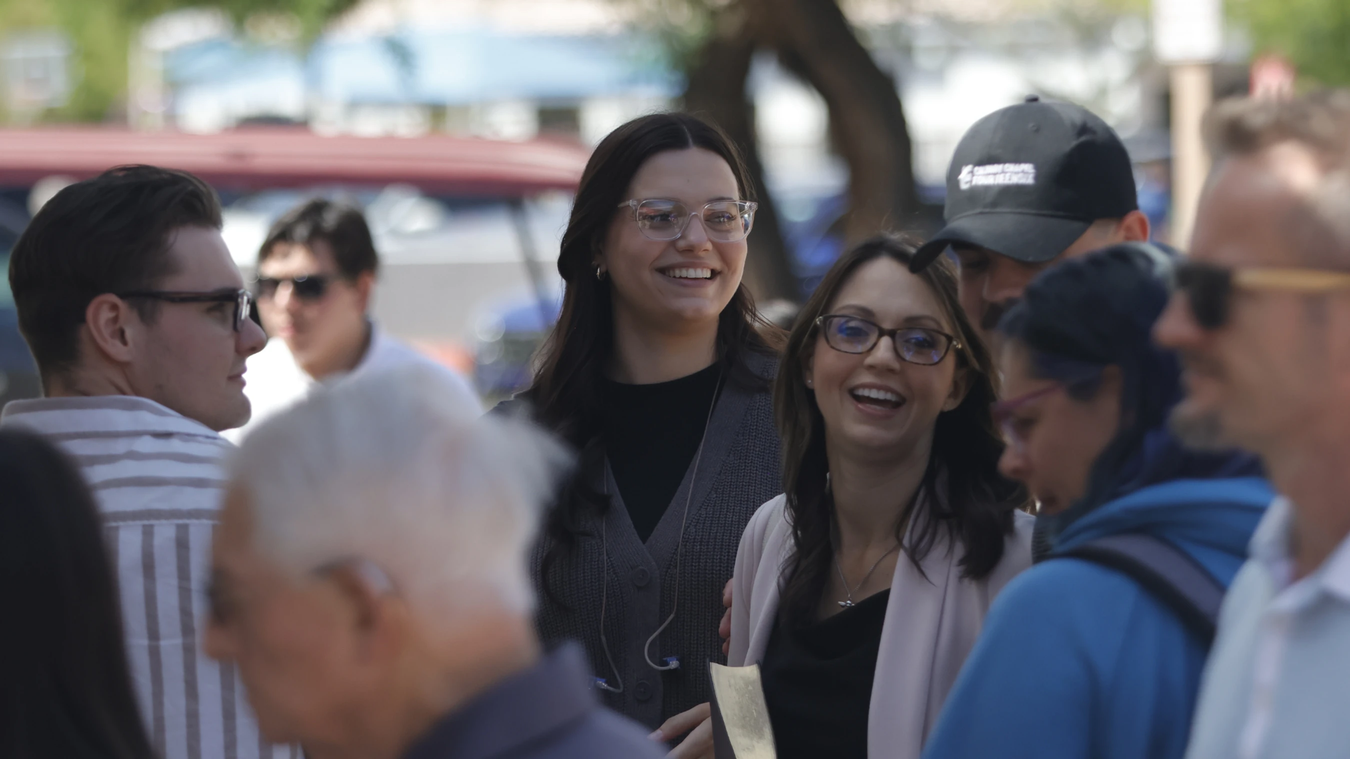 A group of people outdoors, with two women in the center smiling and laughing. One woman in focus wears clear glasses and a grey cardigan.