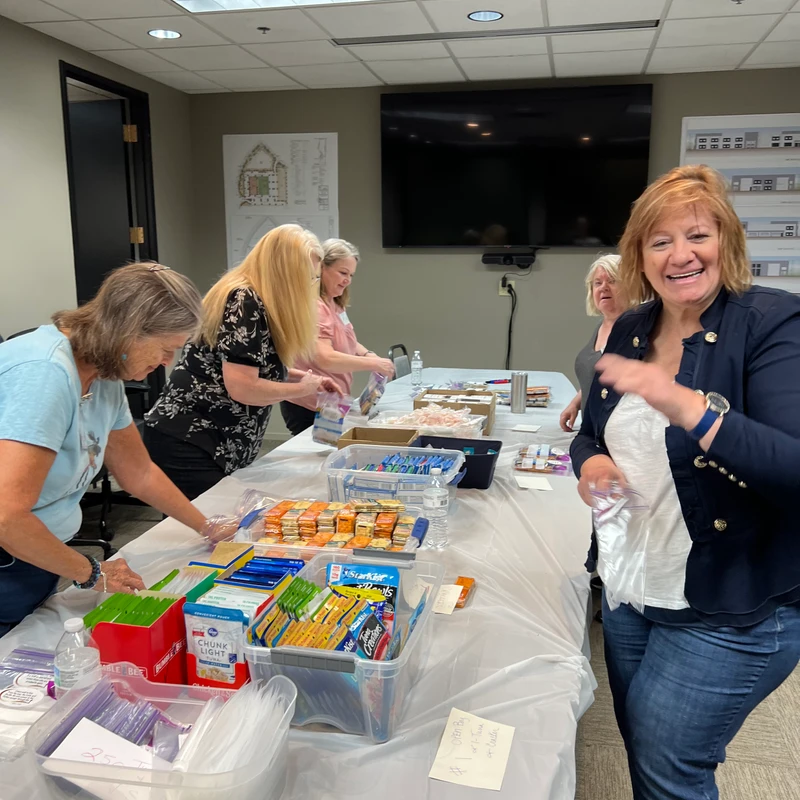 A group of women volunteers smile as they work together in an office, packing snacks and food items into bags on a long table for a charitable cause.