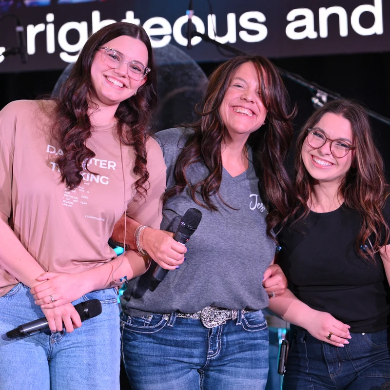 Three women stand close together on a stage, smiling warmly at the camera while holding microphones. A large screen with text is visible in the background.
